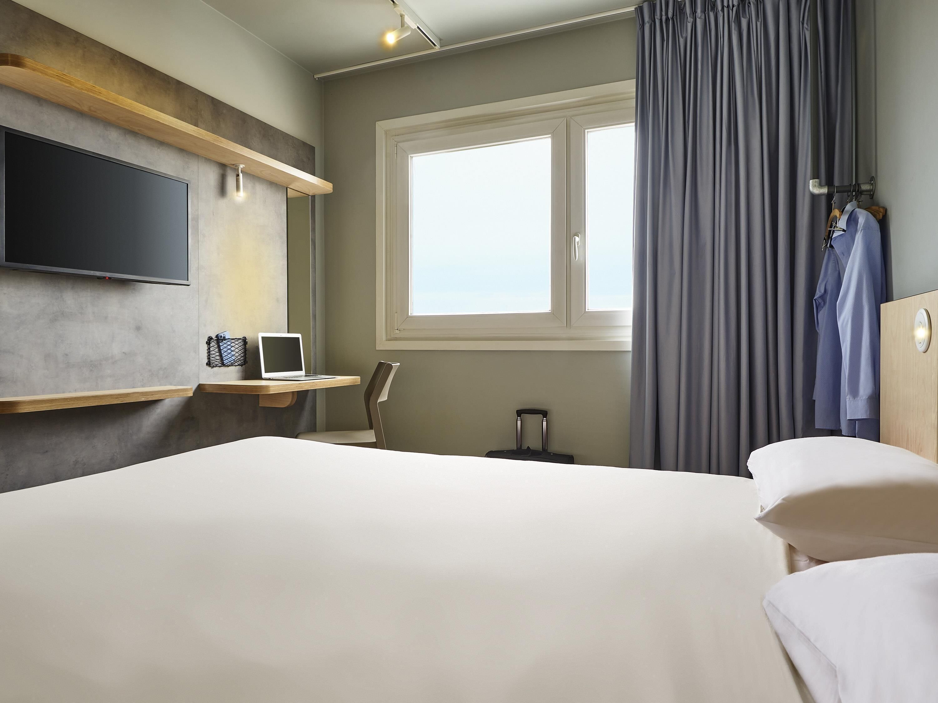 A hotel room with a bed, a desk with a laptop, a TV, a window with curtains, and a suitcase.
