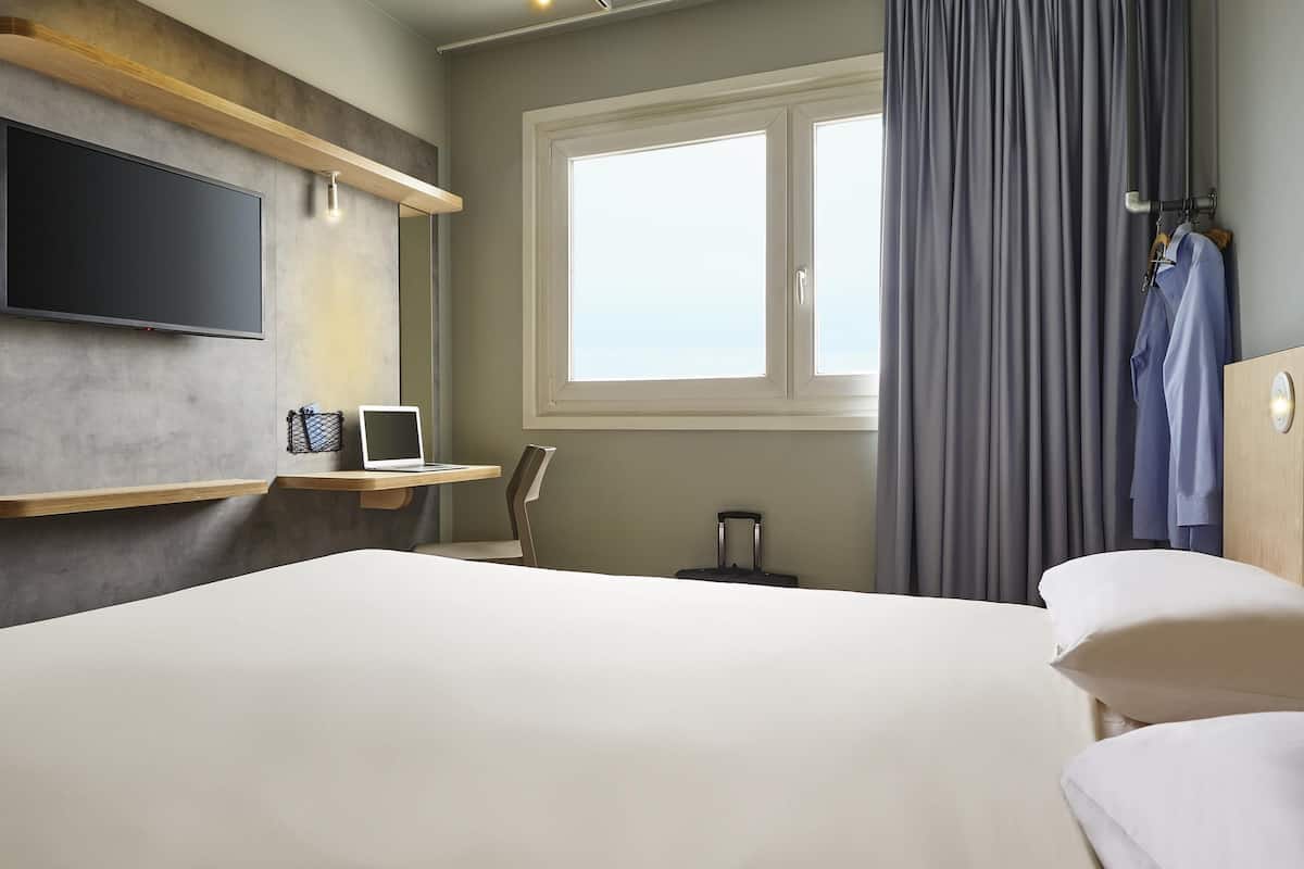 A hotel room with a bed, a desk with a laptop, a TV, a window with curtains, and a suitcase.