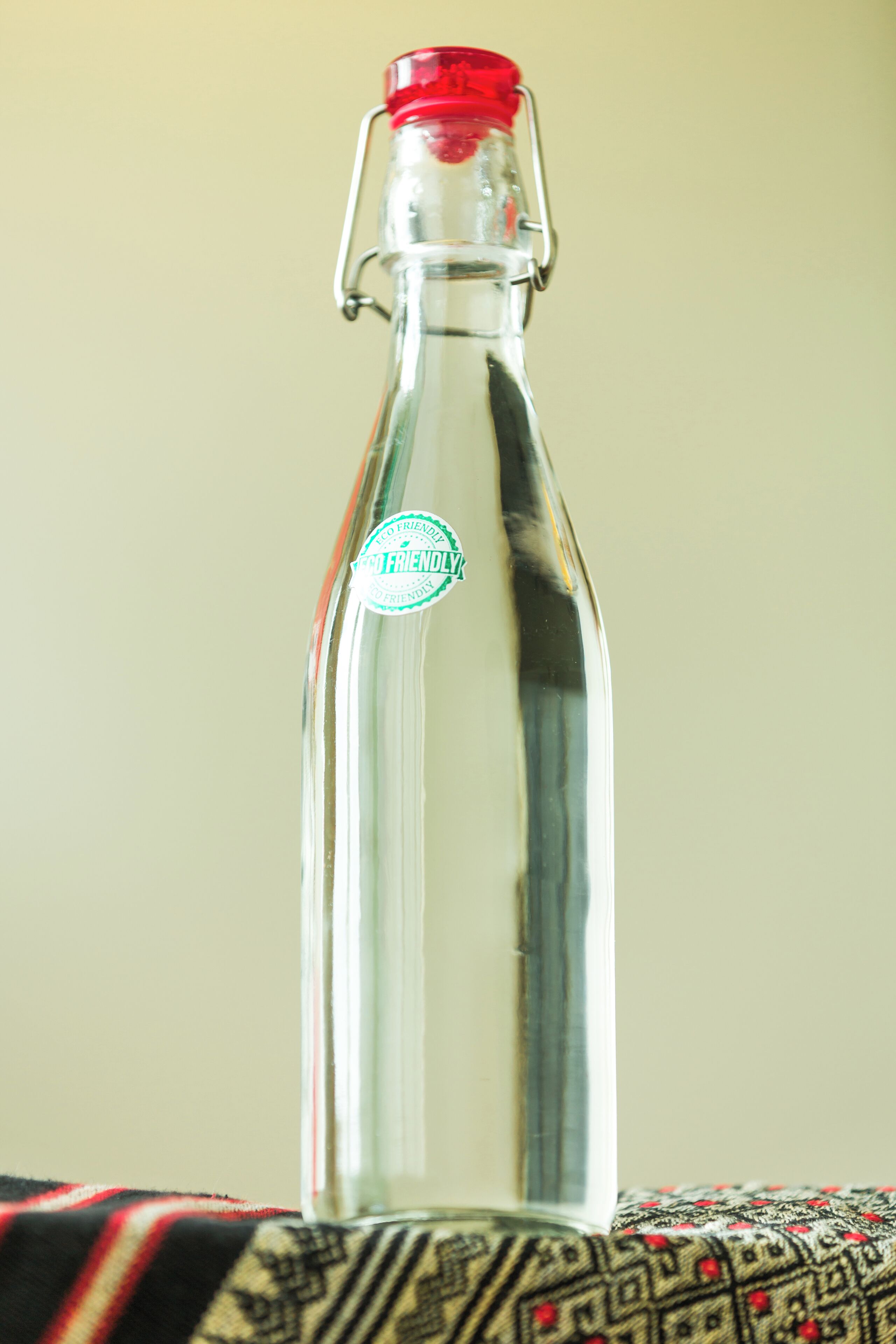 Une bouteille en verre transparent, avec un bouchon rouge et une attache métallique, posée sur un tissu à motifs.