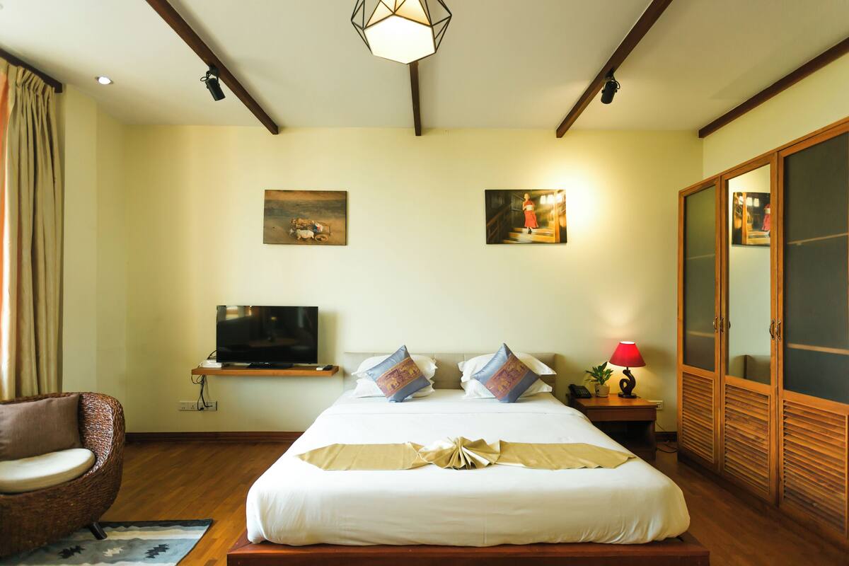 A hotel room with a large bed, a television, a chair, a desk with a lamp, and a wardrobe.