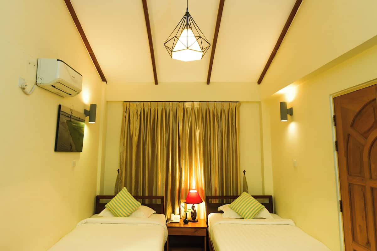 A room with two single beds, wooden flooring, a ceiling with wooden beams, an air conditioning unit, and a hanging light fixture.