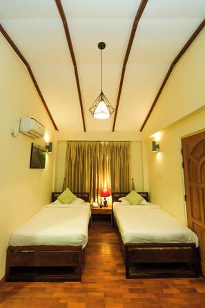 A room with two single beds, wooden flooring, a ceiling with wooden beams, an air conditioning unit, and a hanging light fixture. - Ambassador Hill (Yangon)