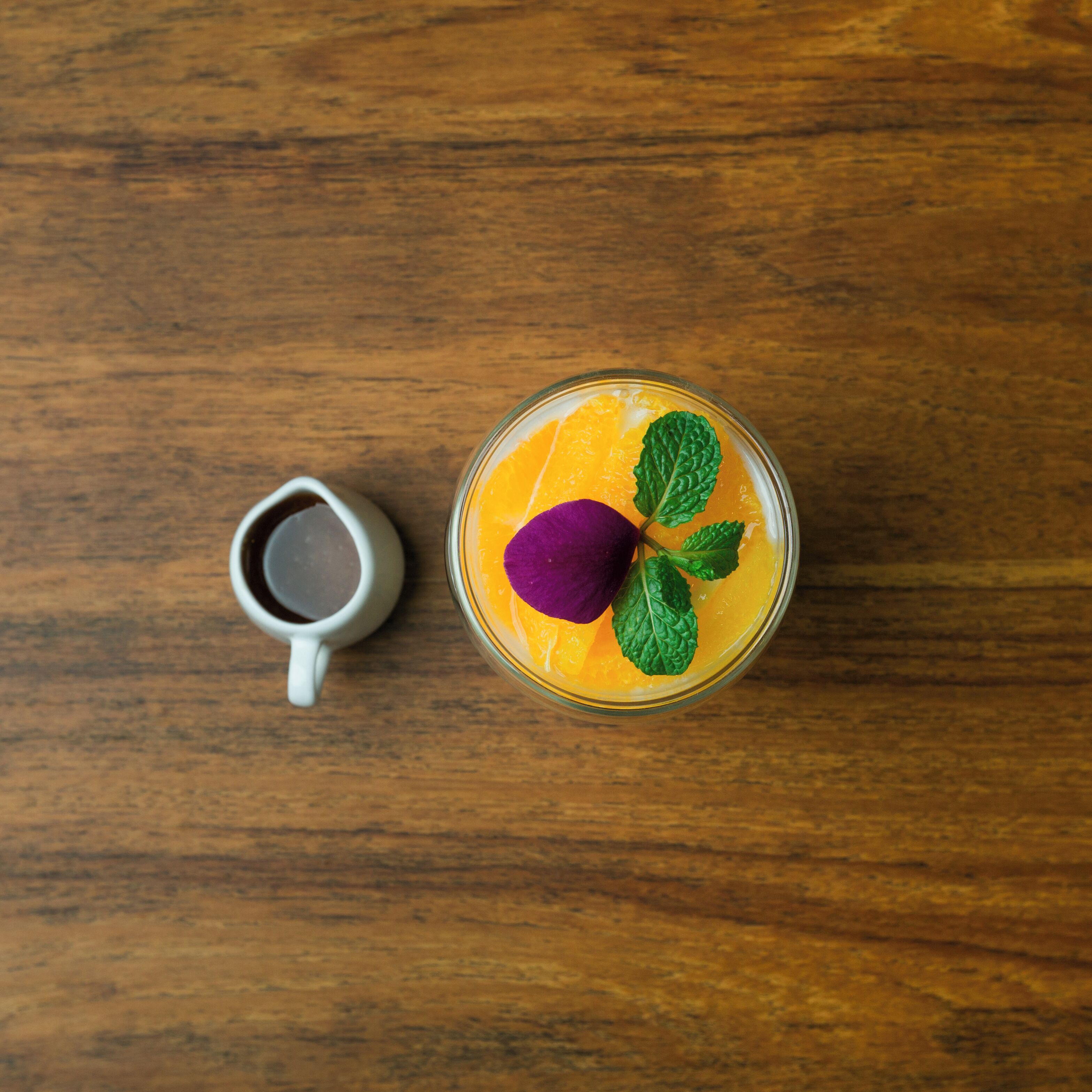 A glass of orange juice with a purple flower and mint leaves on a wooden table next to a white mug.