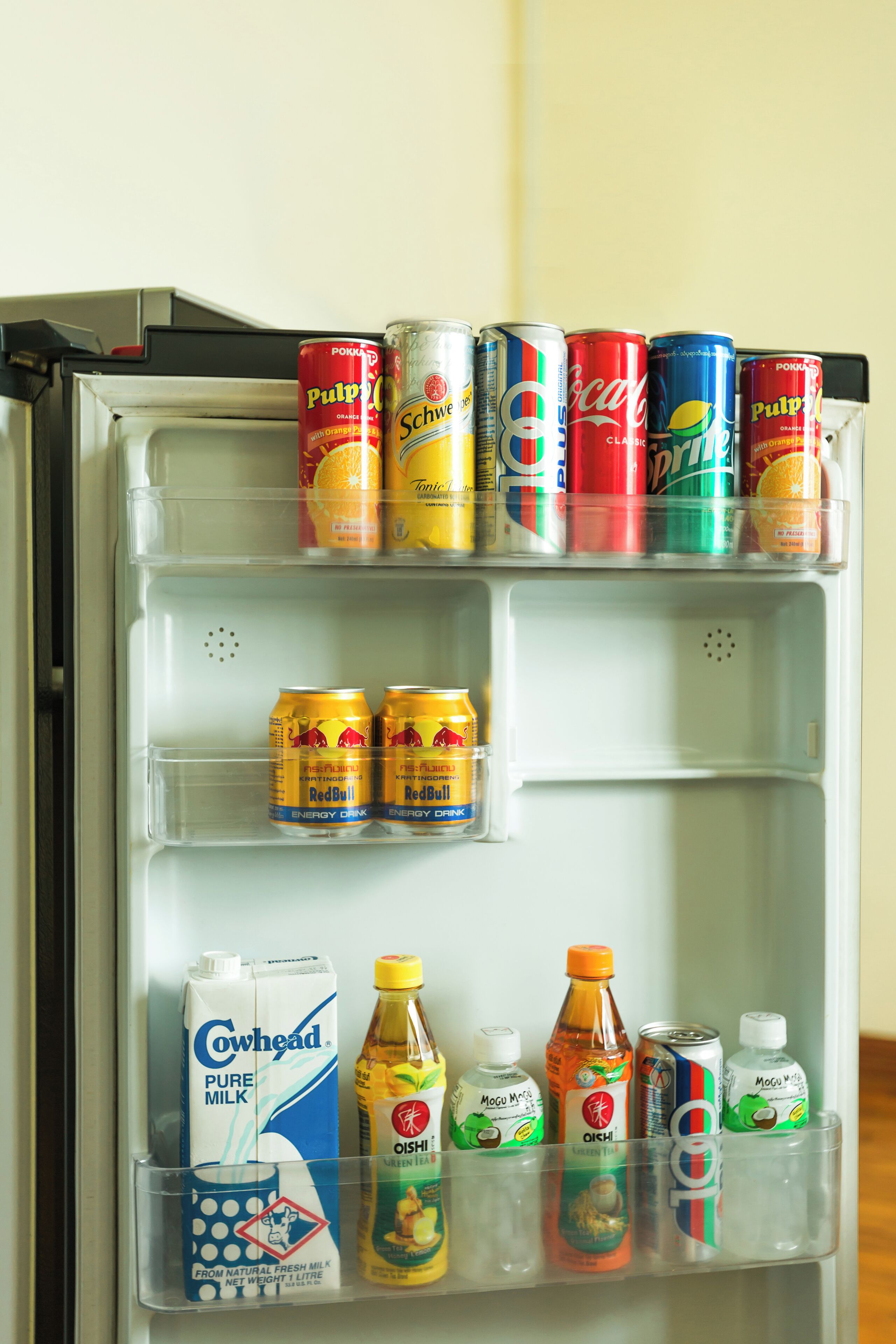 A refrigerator stocked with various beverages including cans of Coca-Cola, Pulp Pops, and Red Bull, as well as cartons of Cowhead milk and bottles of Oishi and Mogu.