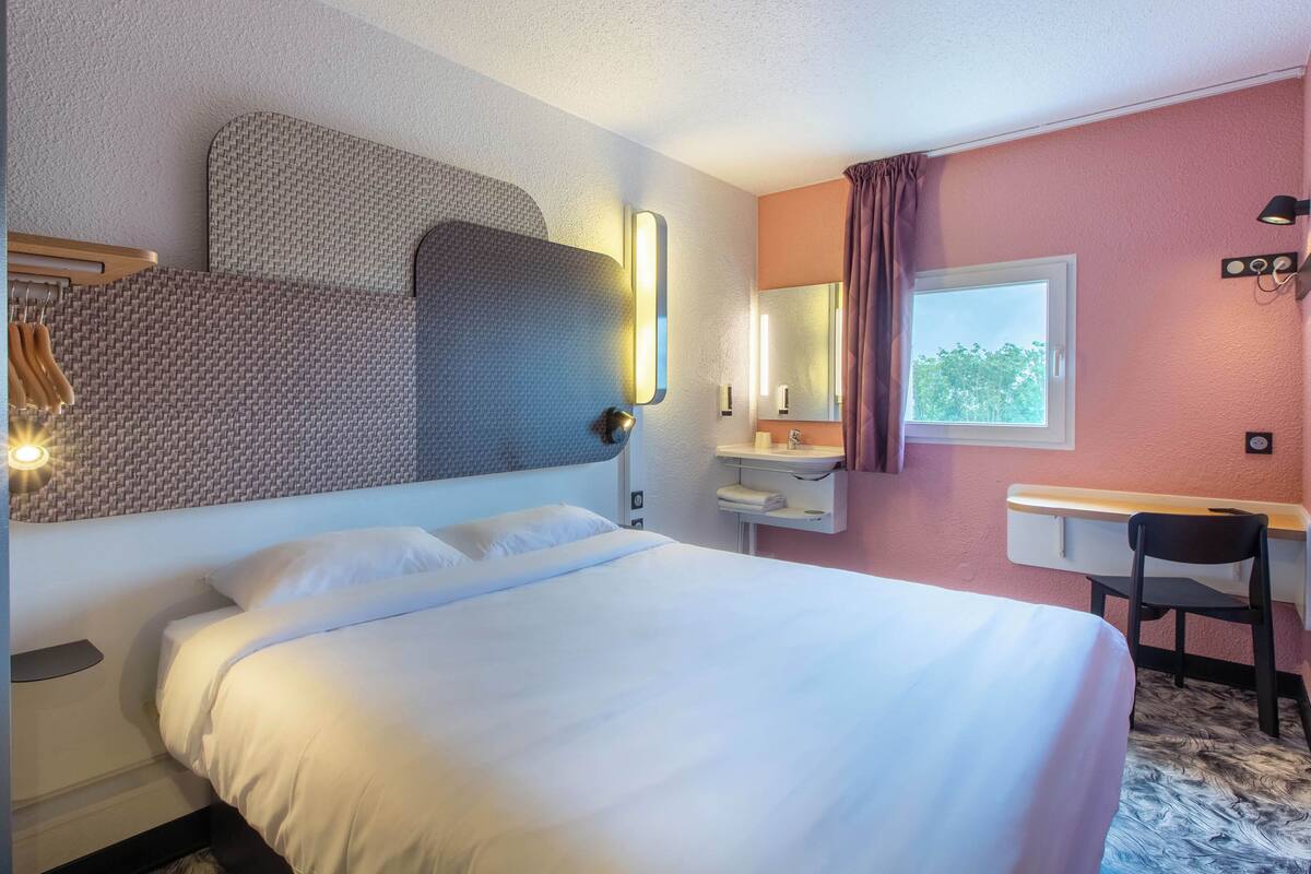 Quarto de hotel com cama, escrivaninha, cadeira, espelho e janela.