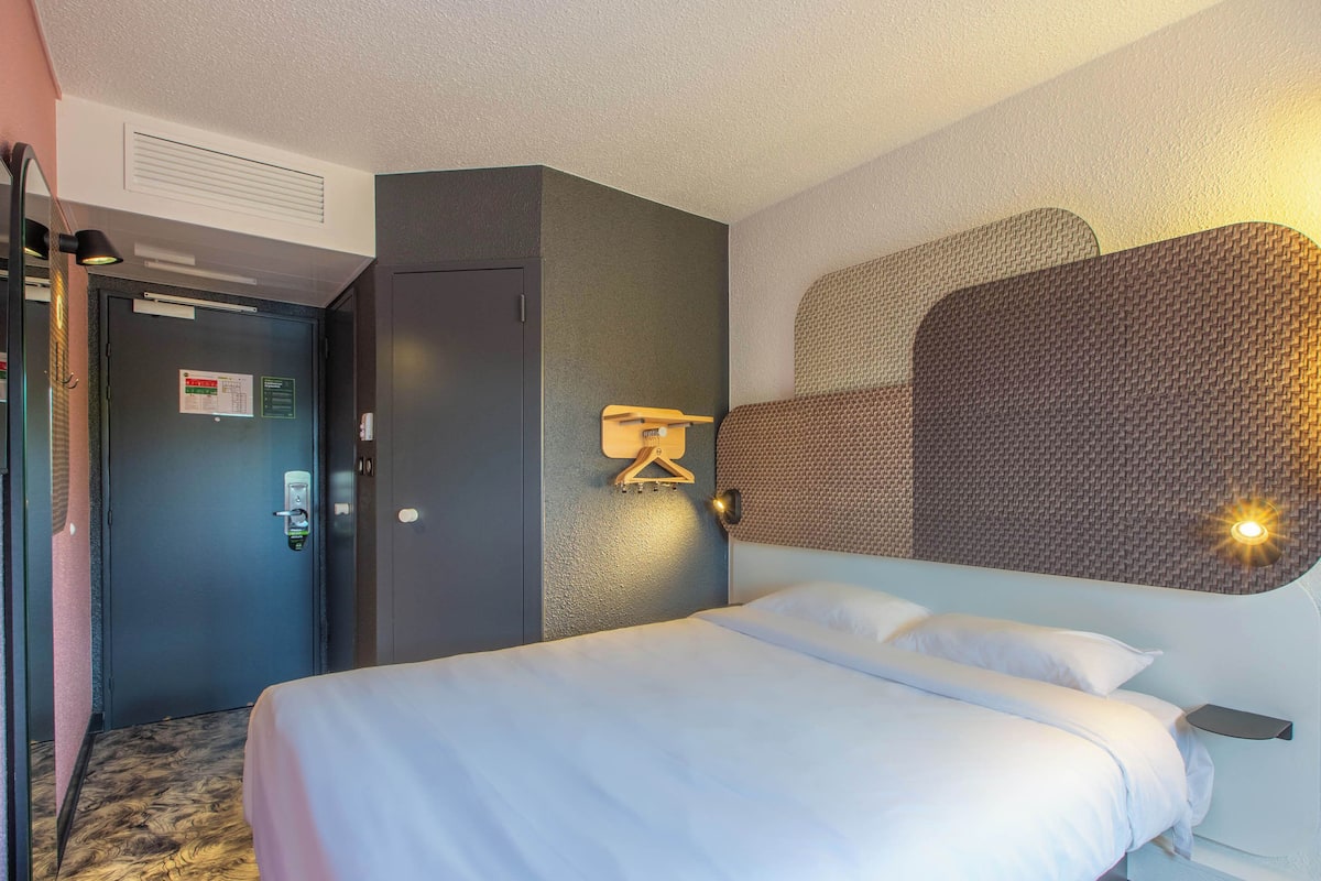 Quarto de hotel com cama, guarda-roupa, porta com fechadura e luminária fixada na parede.