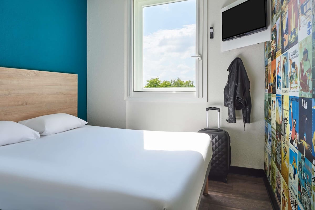 A hotel room with a bed, a window, a TV, and a wall with travel posters.
