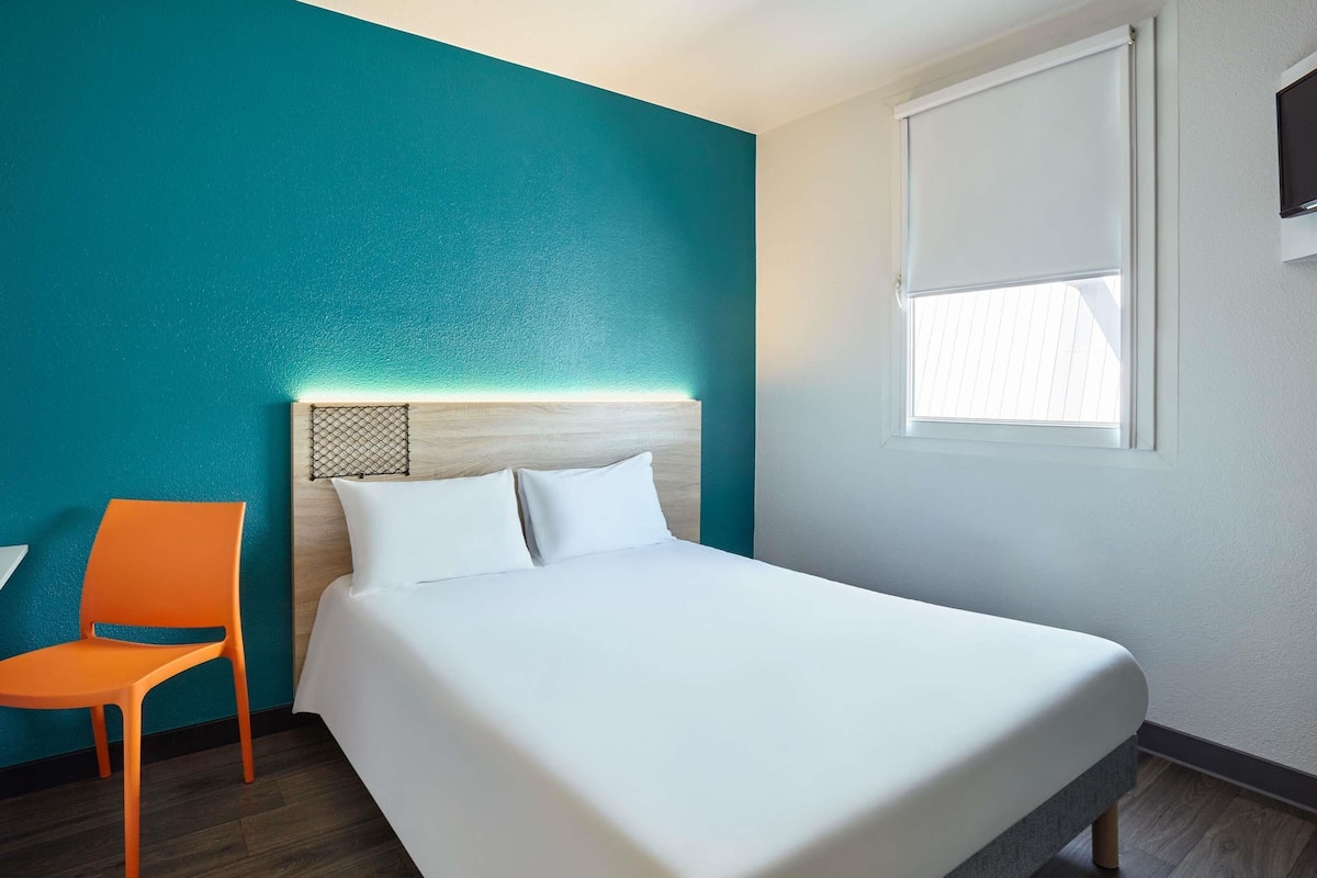 A hotel room with a teal accent wall, a bed with white linens, an orange chair, and a window with blinds.