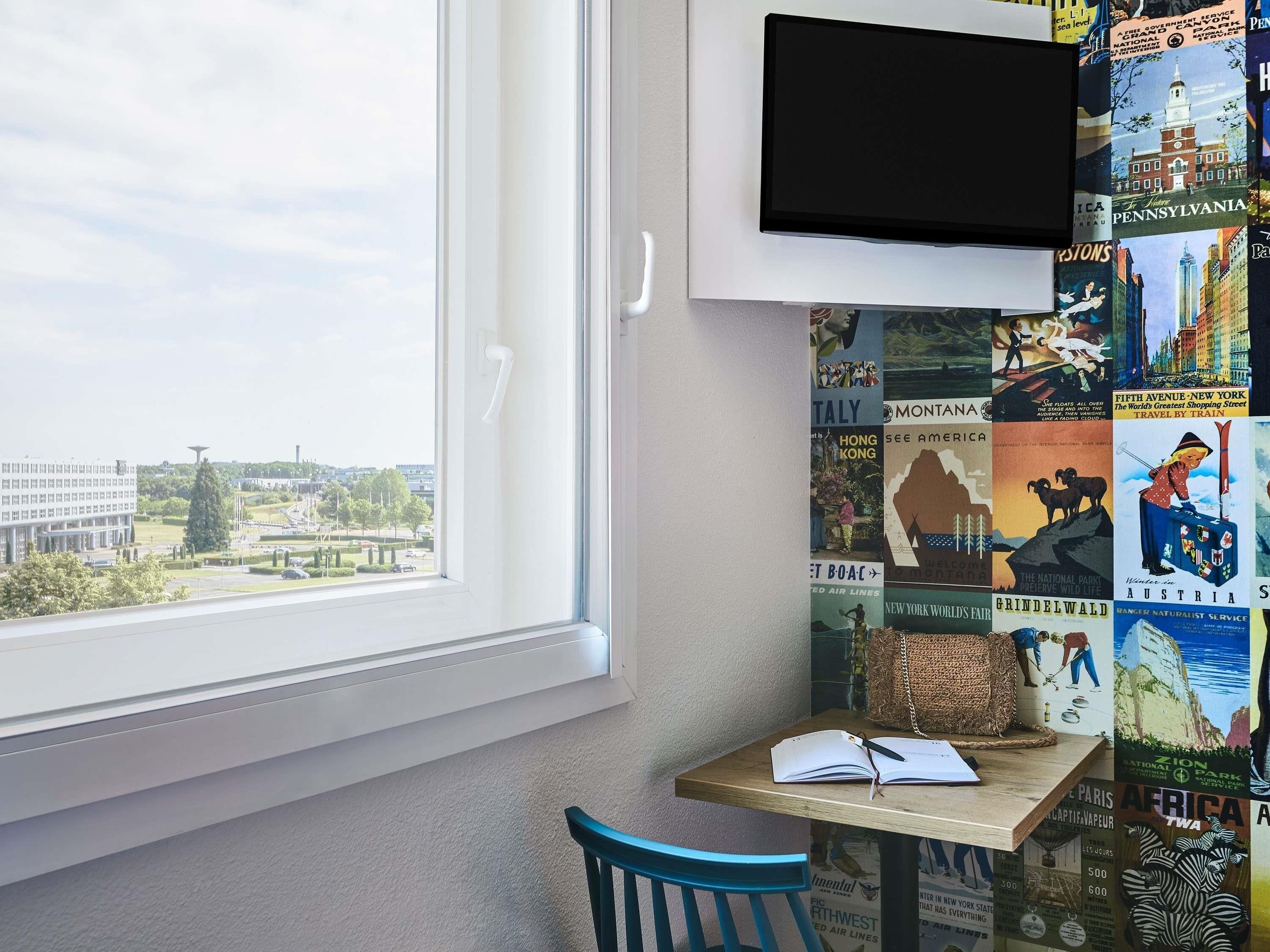 A room with a large window, a flat-screen TV mounted on the wall, a table with a book, and a chair.
