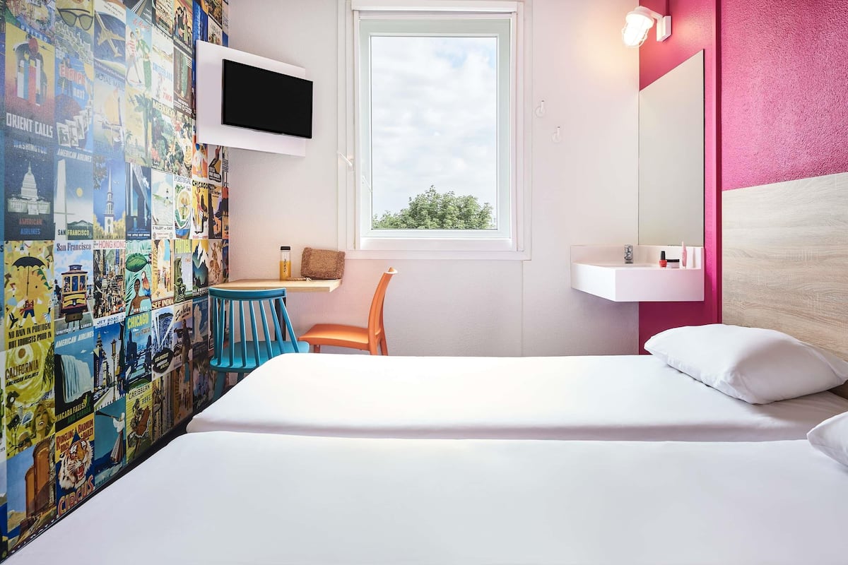 A bedroom with a bed, a TV mounted on the wall, a window, a bedside table, a chair, and a wall covered with travel posters.