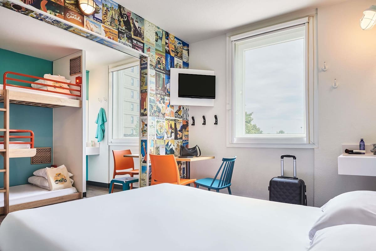 A hotel room with a bunk bed, a desk with a chair, a television, a suitcase, and a window with a view.