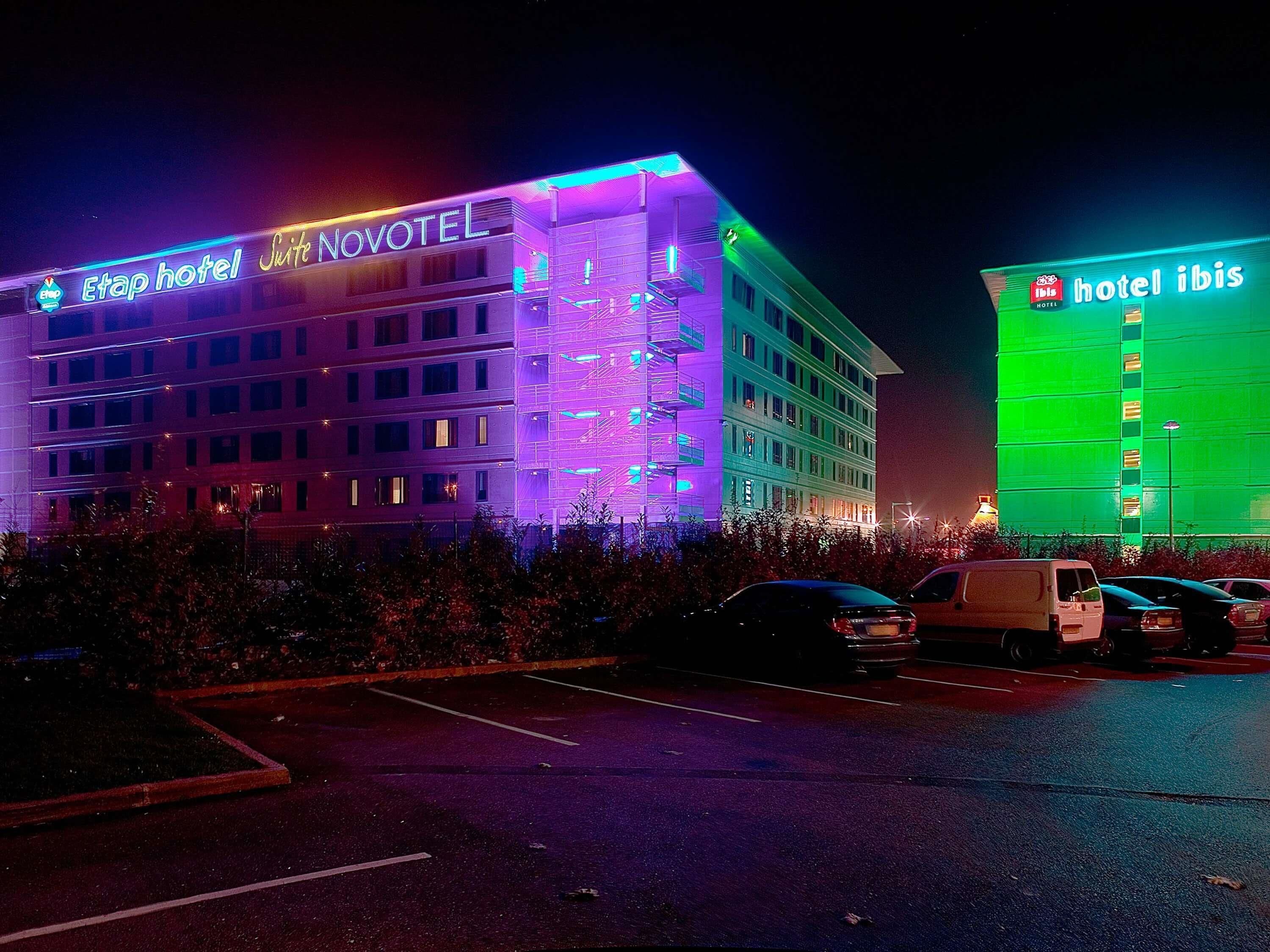 Un hotel con fachada moderna, varios pisos y un estacionamiento con varios vehículos.