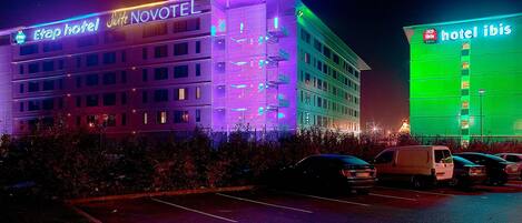 Un hotel con fachada moderna, varios pisos y un estacionamiento con varios vehículos.