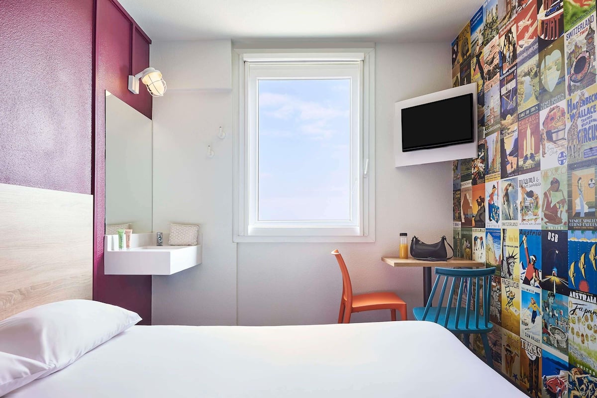 A hotel room with a bed, a window, a TV, a small table, a chair, and a wall with travel posters.