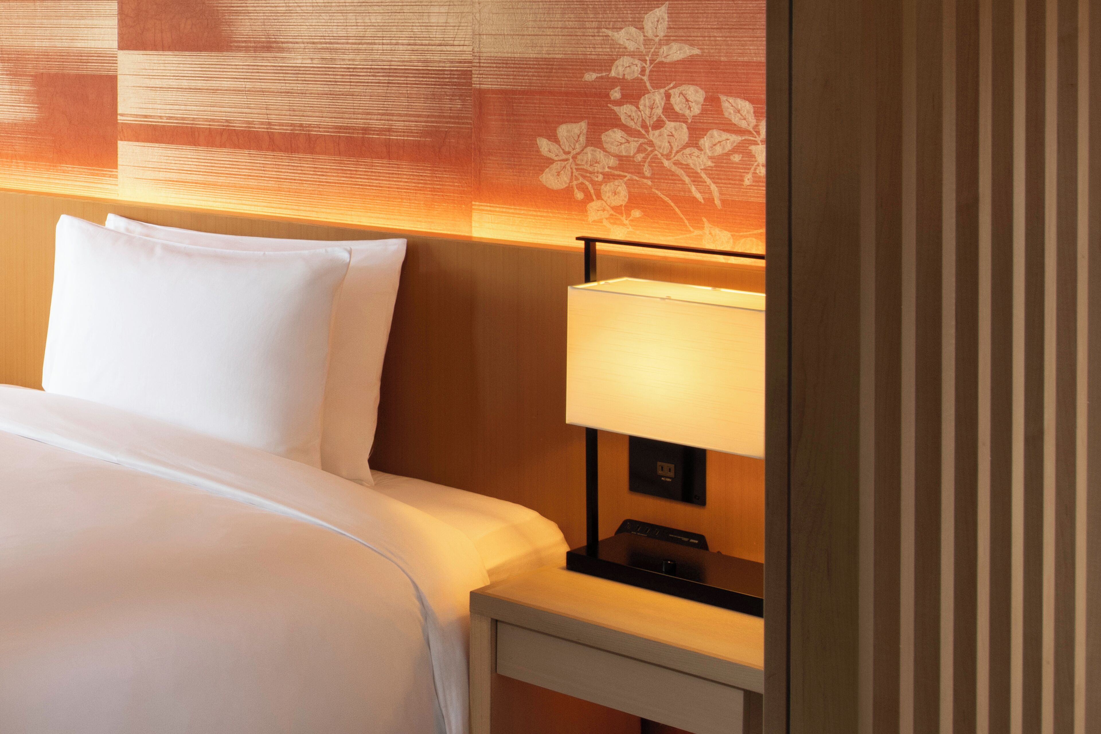 A hotel room with a neatly made bed, a bedside table with a lamp, and a wall with a floral pattern.