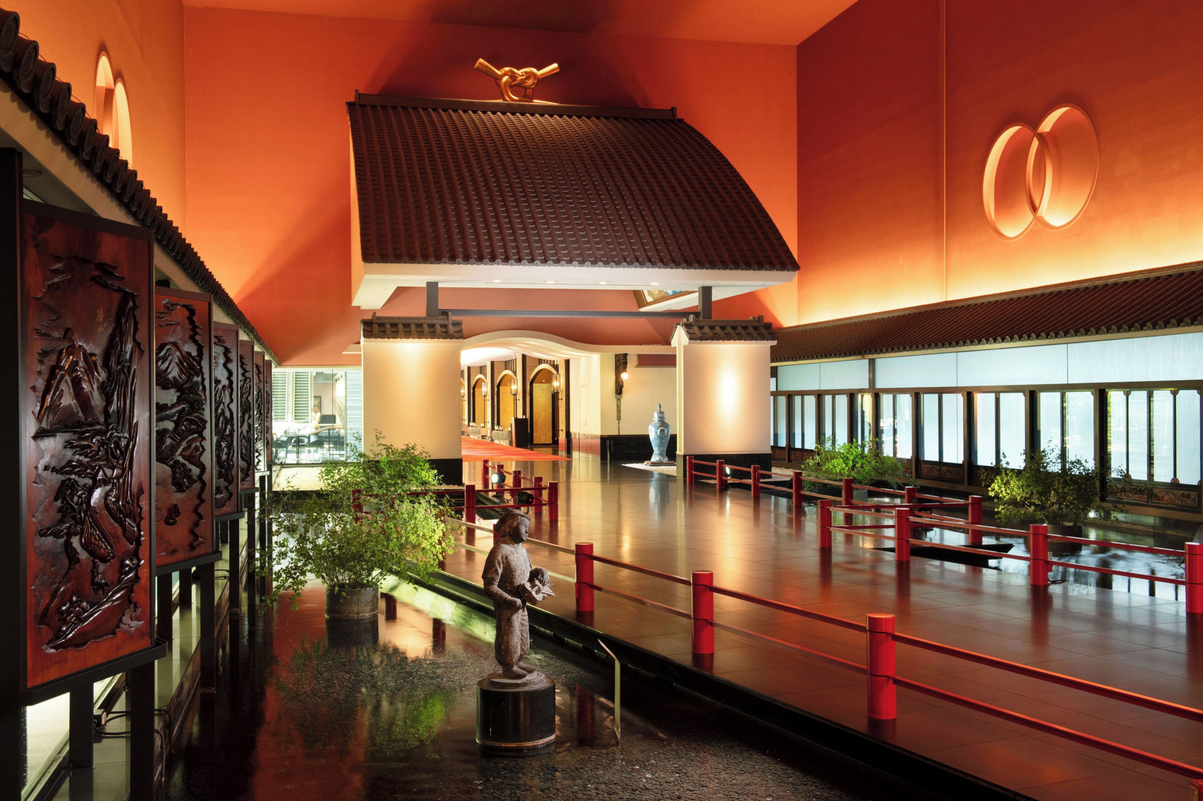 A spacious lobby with a traditional Asian architectural design, featuring a central statue, a red railing, and a large window providing a view of the interior.