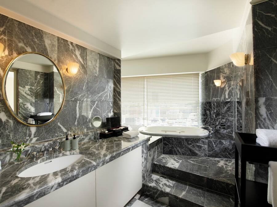 A modern bathroom with marble countertops, a round mirror, a bathtub, and a window with blinds.