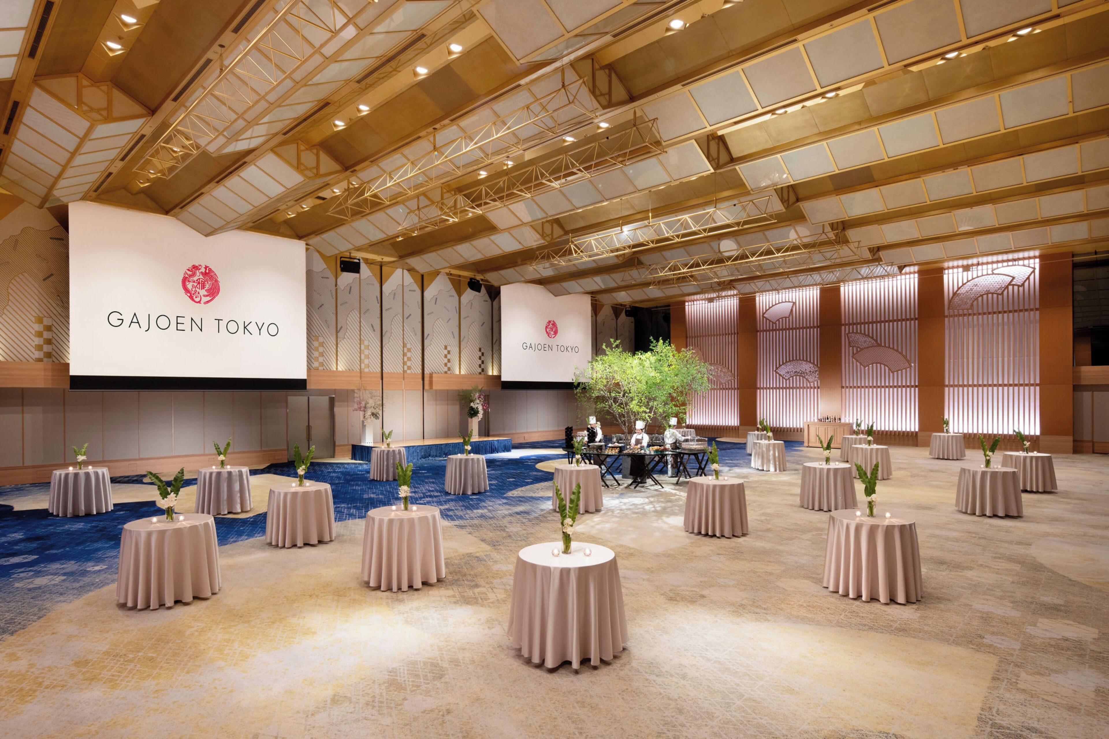 A spacious banquet hall with round tables, a stage, and a ceiling with a geometric design.