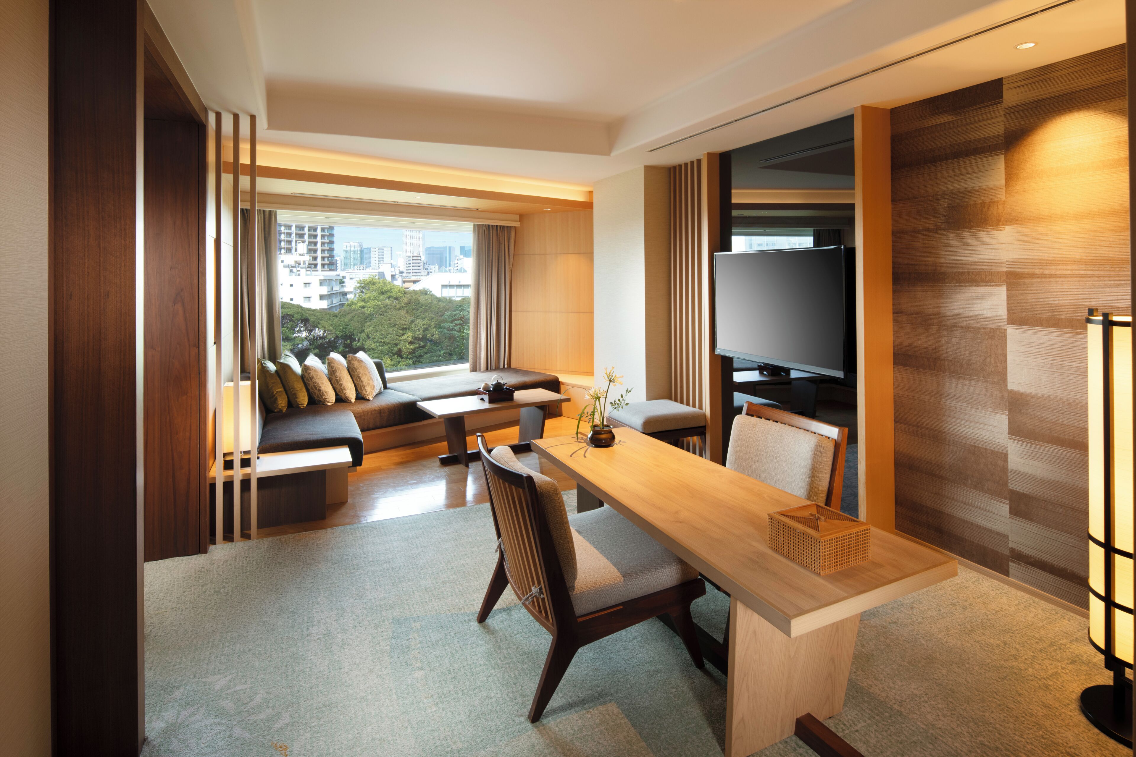 A modern hotel room with a sofa set, a dining table, a TV, and a view of the cityscape.