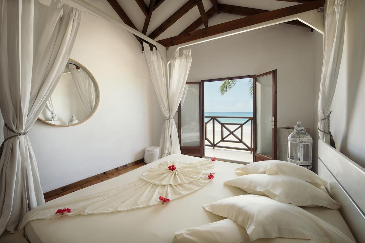 A bedroom with a bed, white curtains, a round mirror, a wooden ceiling, and a view of the sea.