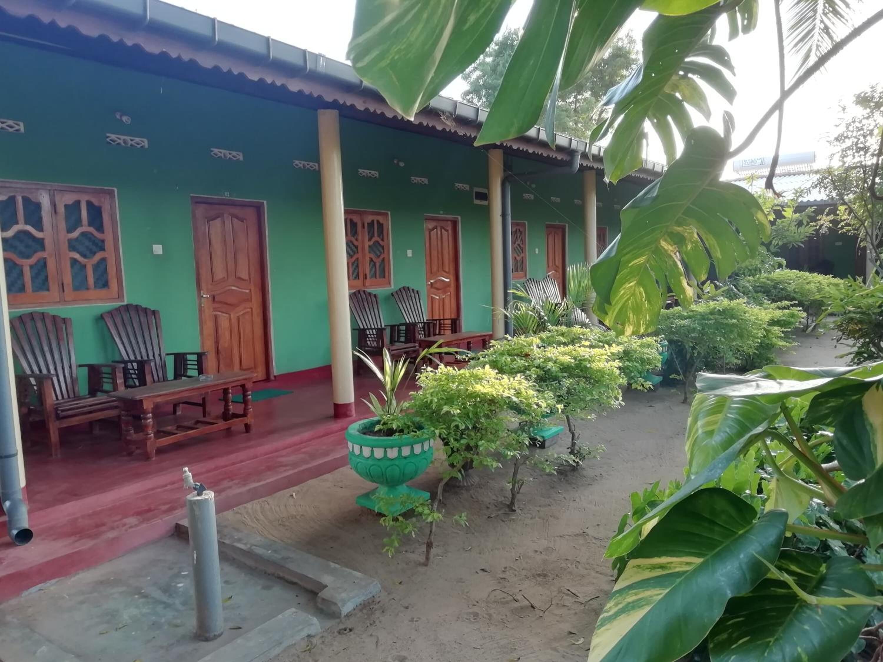 A green building with wooden doors and windows, a covered porch with chairs, and potted plants.