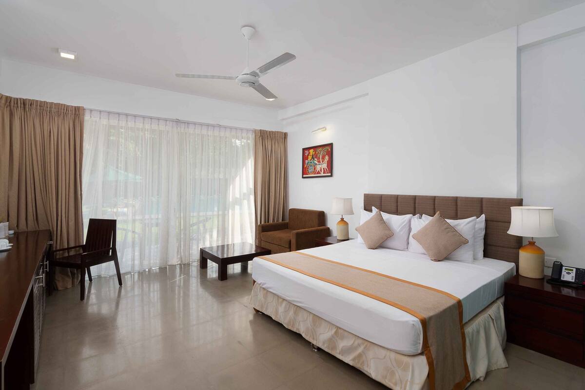 A hotel room with a large bed, a desk, a chair, a sofa, a ceiling fan, and a window with curtains.