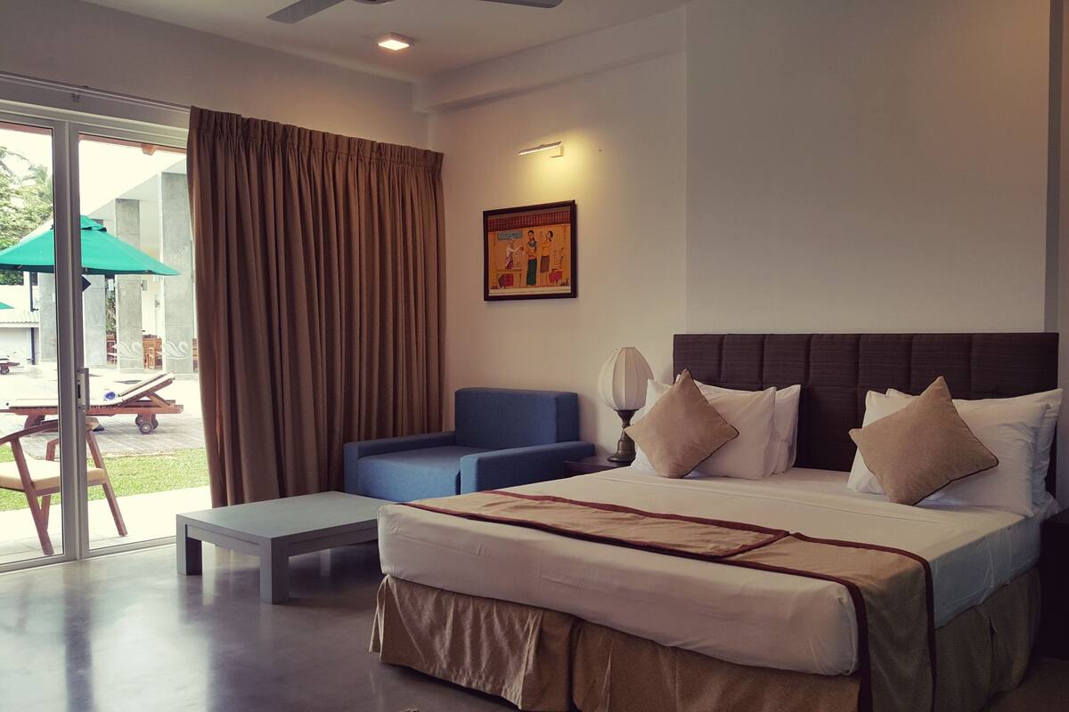 A modern hotel room with a large bed, a blue armchair, a small table, and a view of the outdoor area through a glass door.