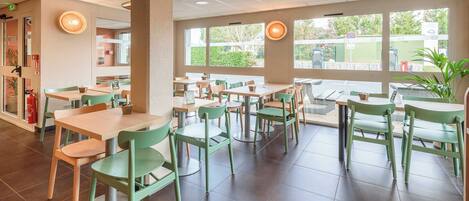 Interior de restaurante con sillas verdes y mesas blancas, ventanas amplias y un techo con diseño de cuadrícula.