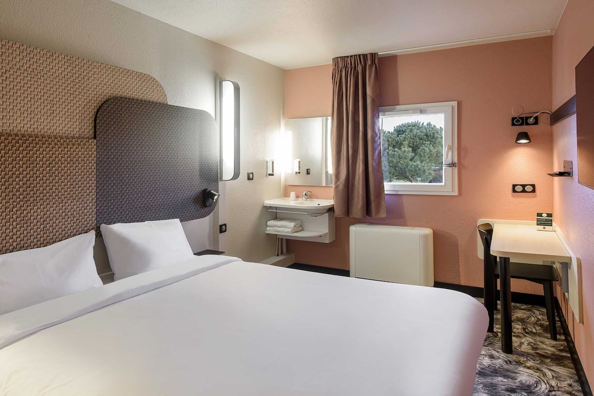 A hotel room with a large bed, a bedside table, a small desk, a chair, a window with a view of trees, a sink, and a mirror.