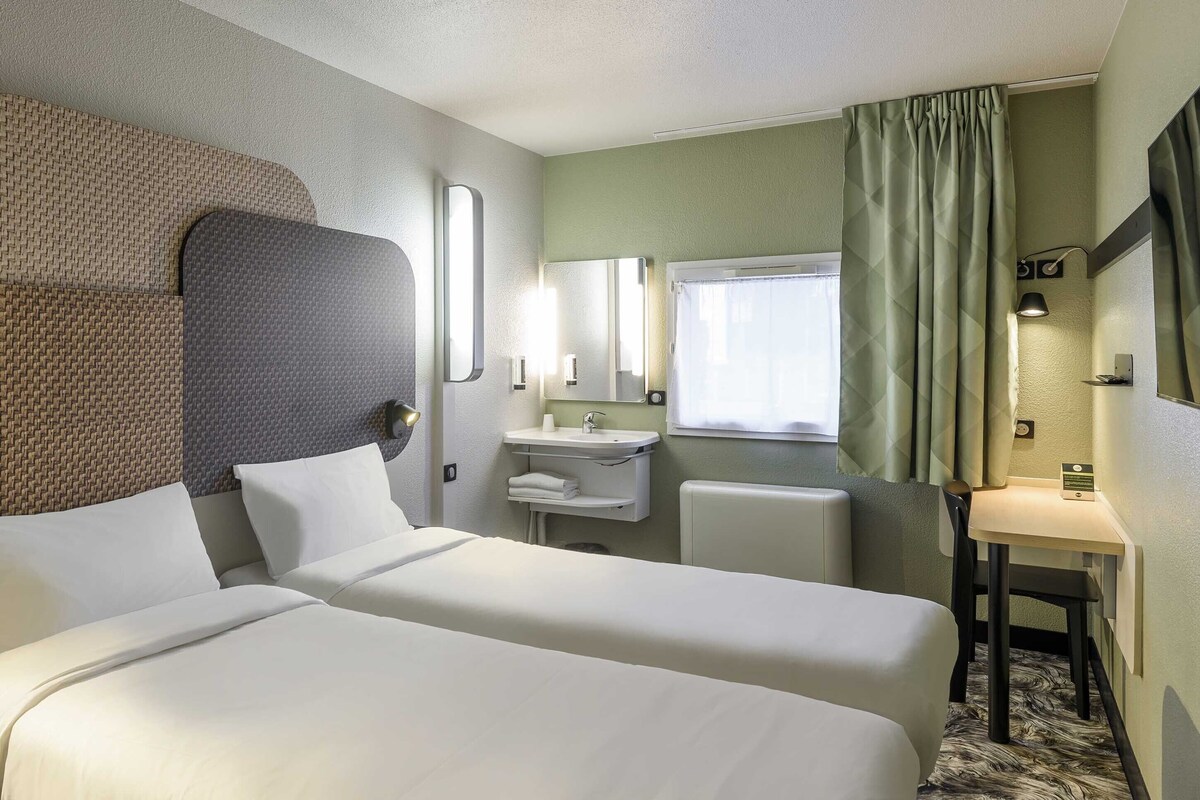 A hotel room with two beds, a bathroom with a sink and mirror, and a small table with a chair.