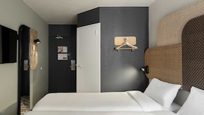 A hotel room with a bed, a wardrobe, a mirror, a wall-mounted light, and a hanging coat rack. - B&B HOTEL Toulouse Cité de l'Espace Hurel (Toulouse)
