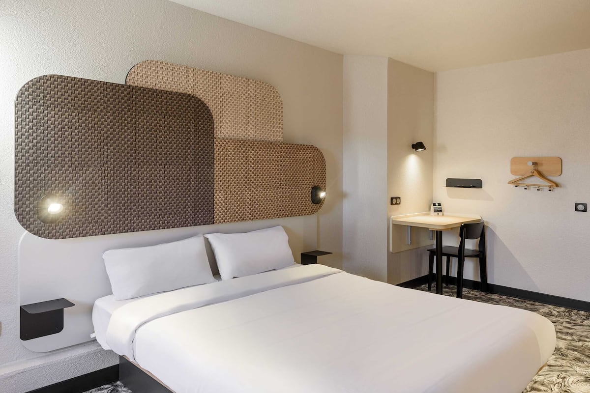 A hotel room with a large bed, a desk with a chair, a mirror, and a wall-mounted coat rack.