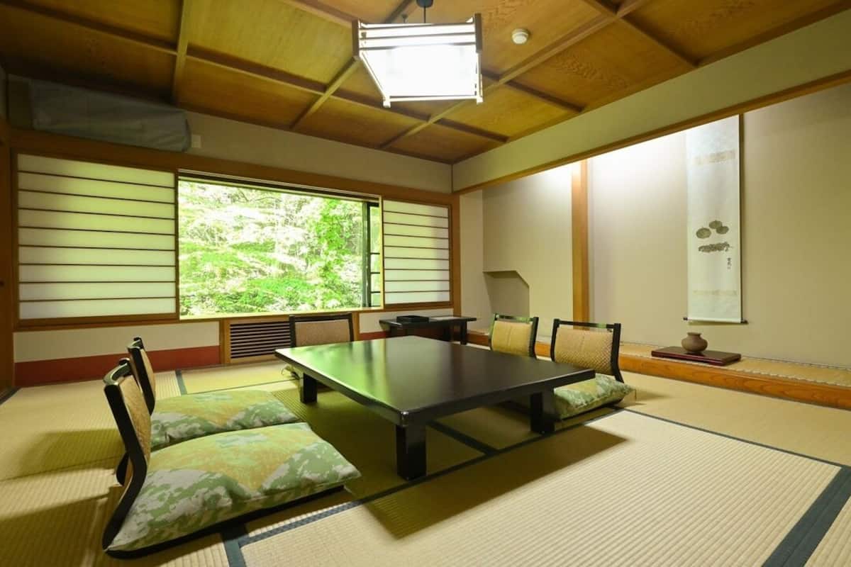 A traditional Japanese room with tatami flooring, a low table, cushions, sliding doors, and a view of greenery.