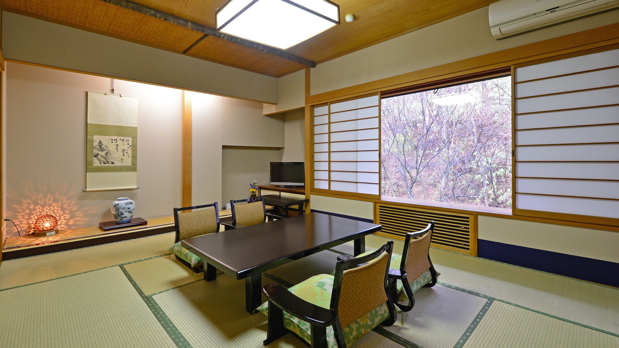 Una habitación japonesa tradicional con suelo de tatami, una mesa baja, sillas, un televisor y un ventanal con puertas shoji.