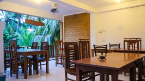 A dining area with wooden tables and chairs, a brick accent wall, and a view of tropical foliage through large windows.