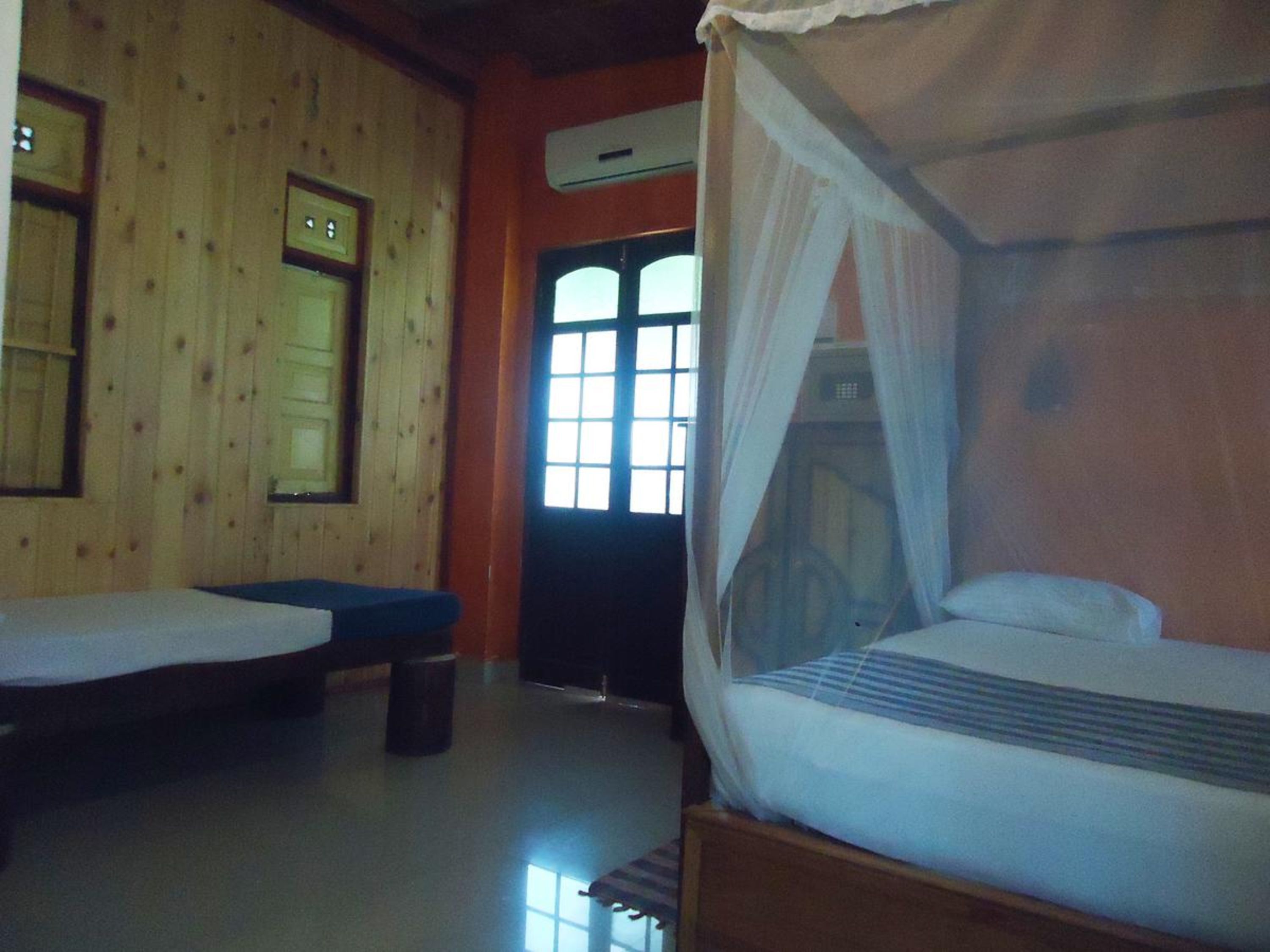 Una habitación con dos camas, una con dosel, paredes de madera, un ventanal y un equipo de aire acondicionado.