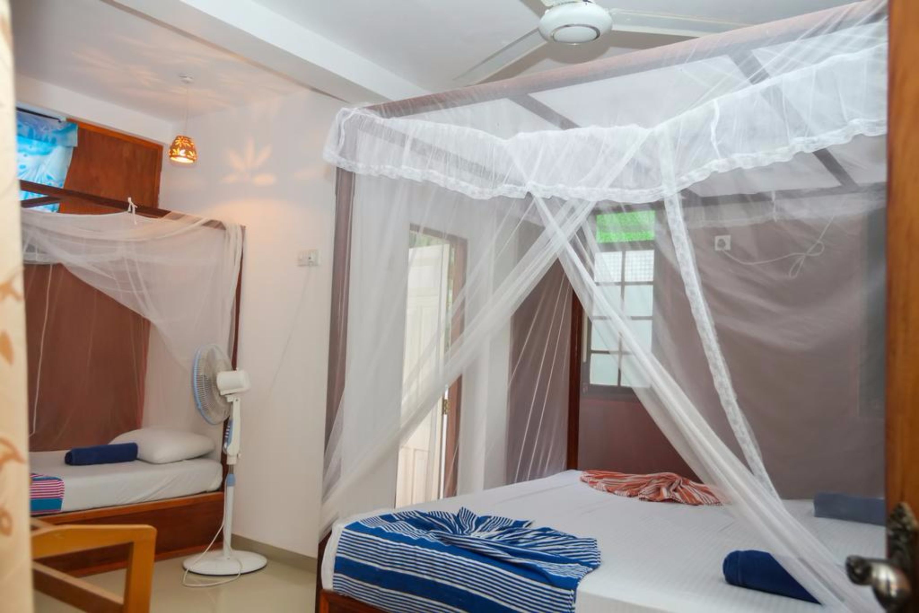 A bedroom with a bed under a mosquito net, a bedside fan, and a window with curtains.