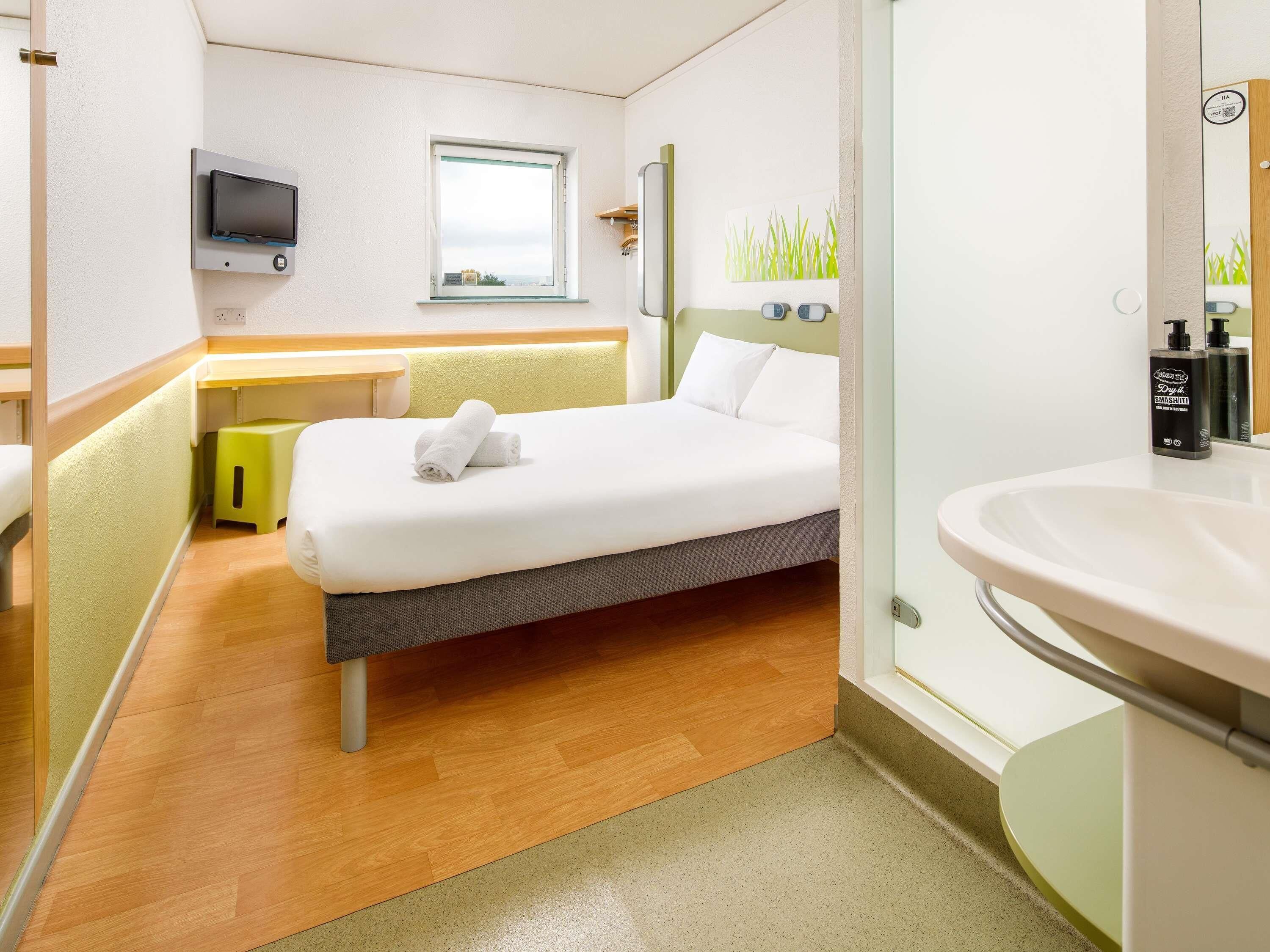 Une chambre d’hôtel moderne équipée d’un lit, d’une table de chevet, d’une télévision, d’une fenêtre et d’une salle de bain avec un lavabo et un miroir.