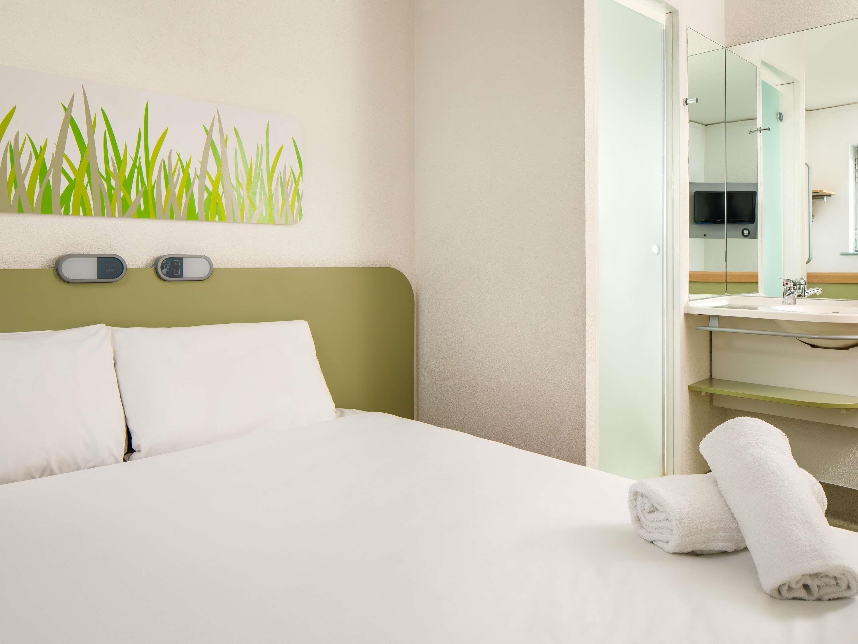 A hotel room with a bed, a wall-mounted digital clock, a bathroom with a sink and mirror, and a towel on the floor.