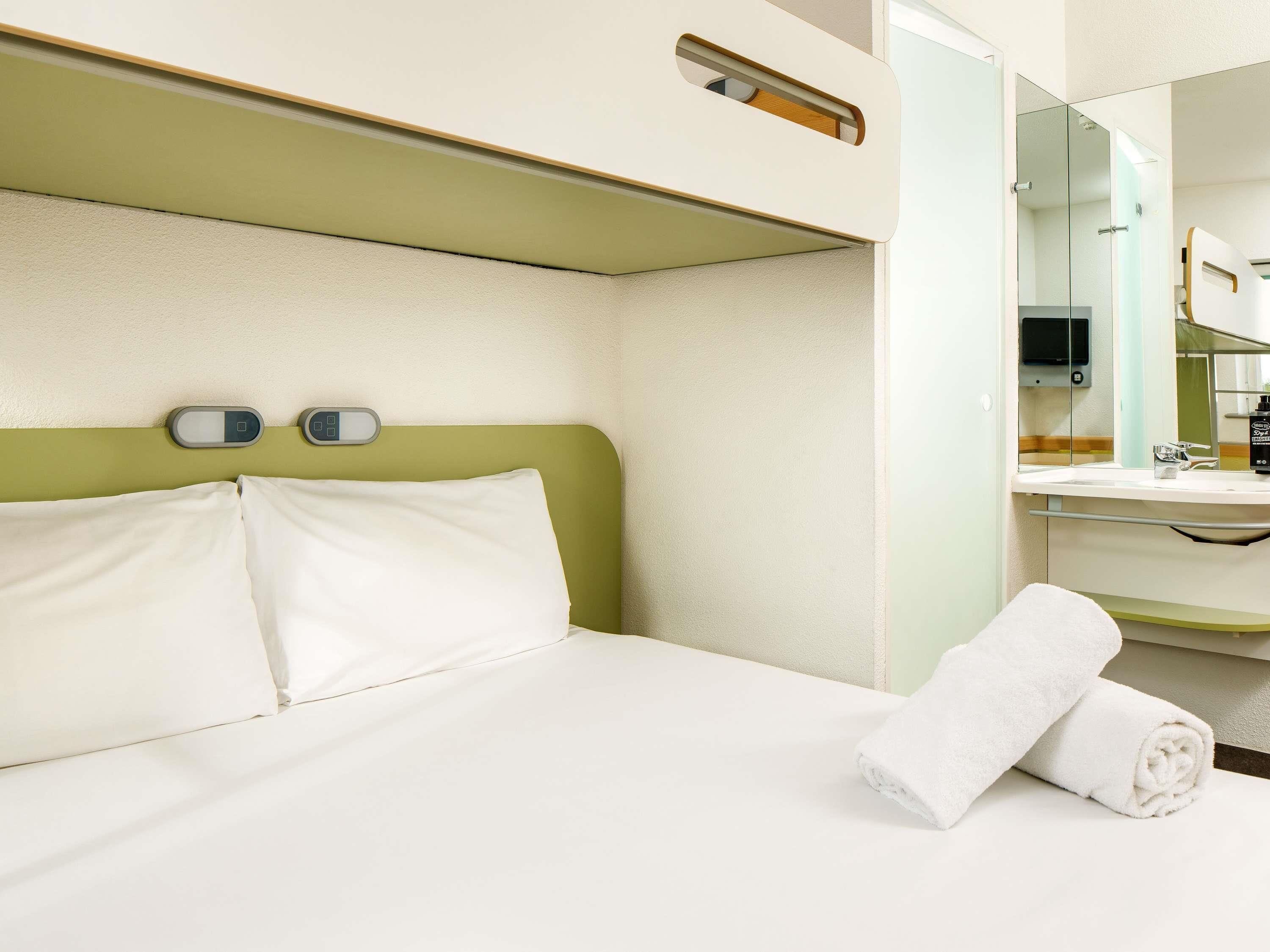 Quarto de hotel com cama, cabeceira verde, mesa de cabeceira com espelho e toalha branca.