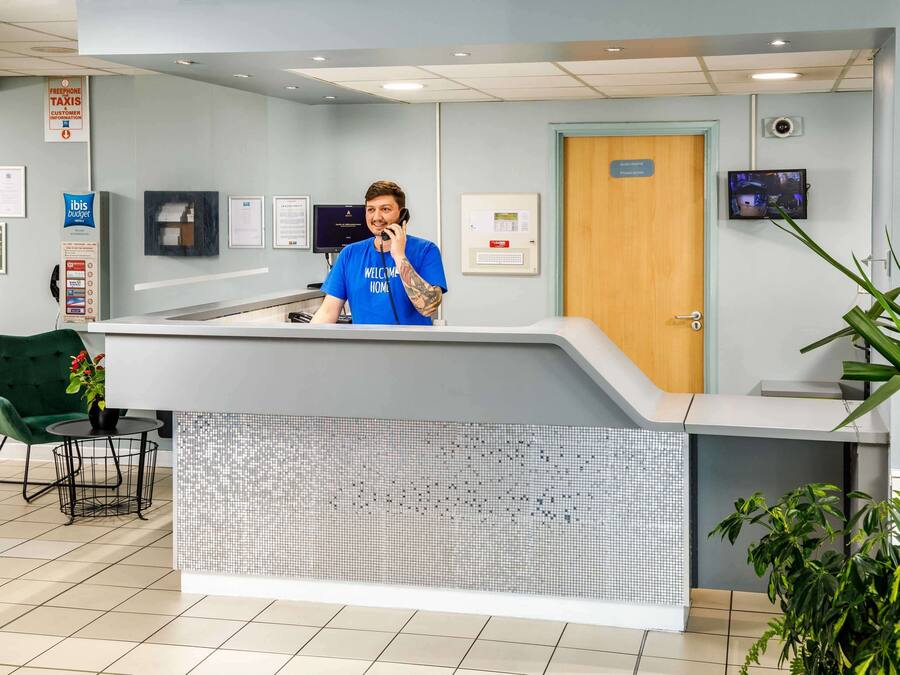 Een receptiebalie met een persoon in een blauw shirt, een beveiligingscamera, een brandmeldinstallatie en een bordje met daarop informatie over gratis taxi's.