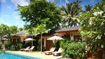 Zona junto a la piscina con sillones de descanso y sombrillas, rodeada de vegetación exuberante y palmeras.