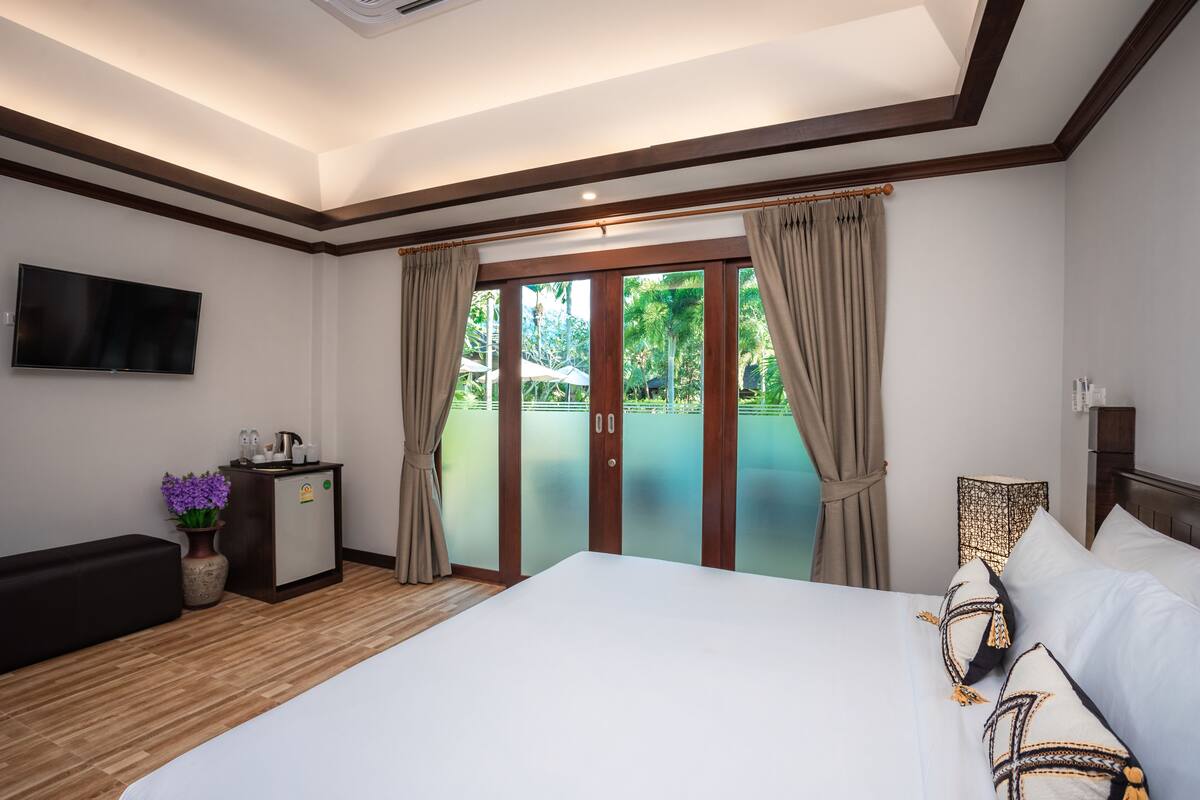 Ein Hotelzimmer mit Bett, Wandfernseher, kleinem Schreibtisch mit Blumenvase und einer Schiebetür zum Balkon.