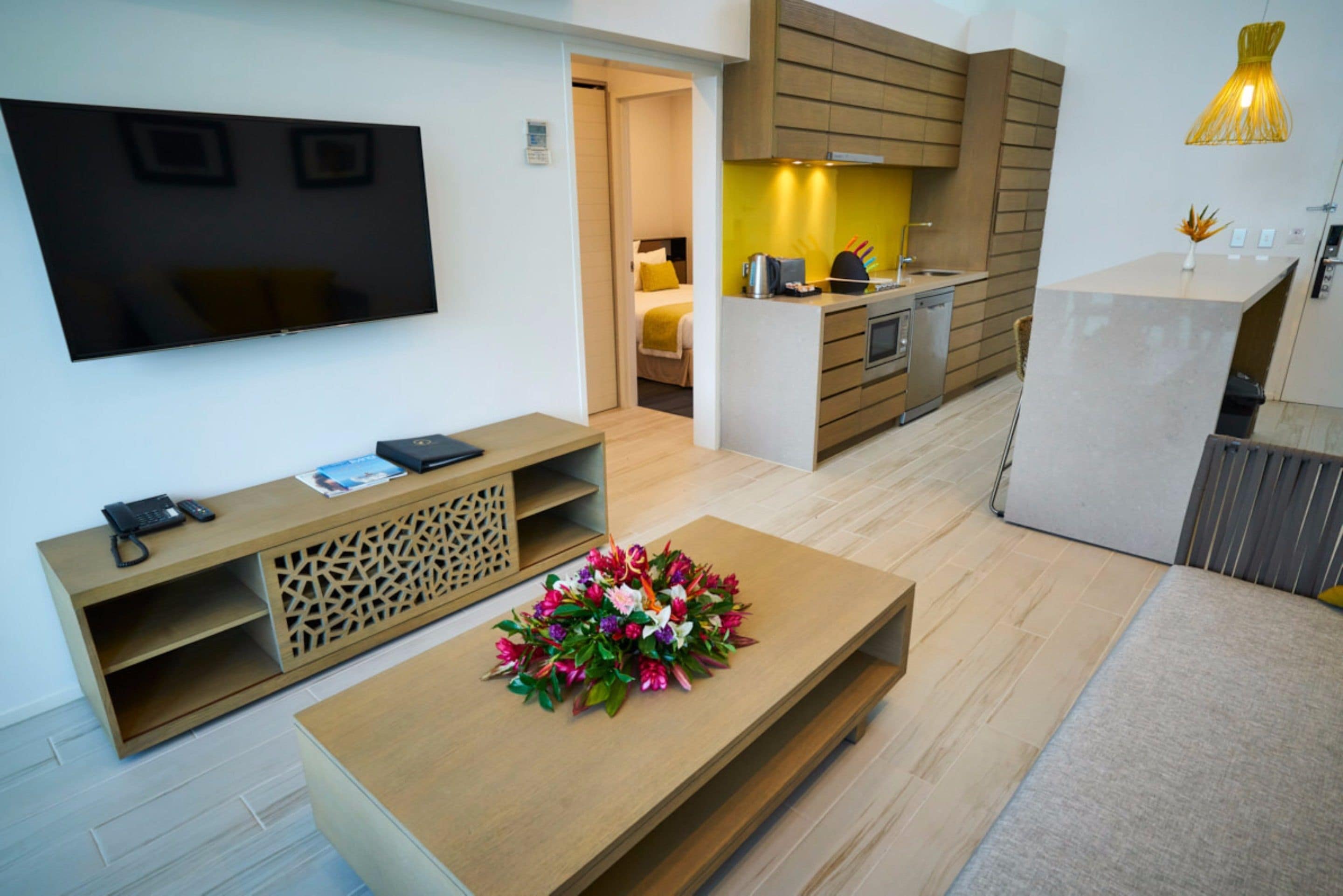 Een moderne hotelkamer met een flatscreen-tv, een houten salontafel met een bloemstuk erop, een kitchenette en een bed met een geel hoofdeinde.