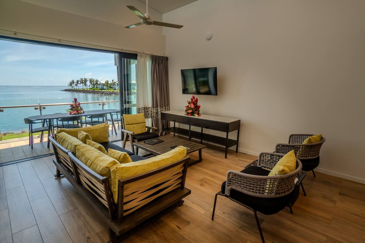 A spacious living area with a wooden floor, a sofa set, a dining table, and chairs. A flat-screen TV is mounted on the wall, and a ceiling fan is visible.