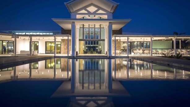 Un hotel moderno con un atrio central, una piscina en primer plano y varias zonas de restauración visibles a través de la fachada de vidrio.
