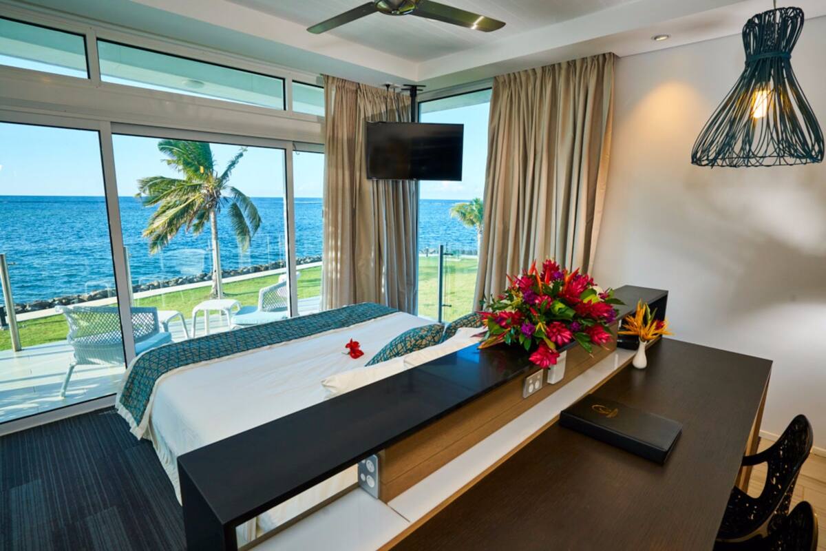 A hotel room with a bed, a desk, a TV, and a view of the ocean.