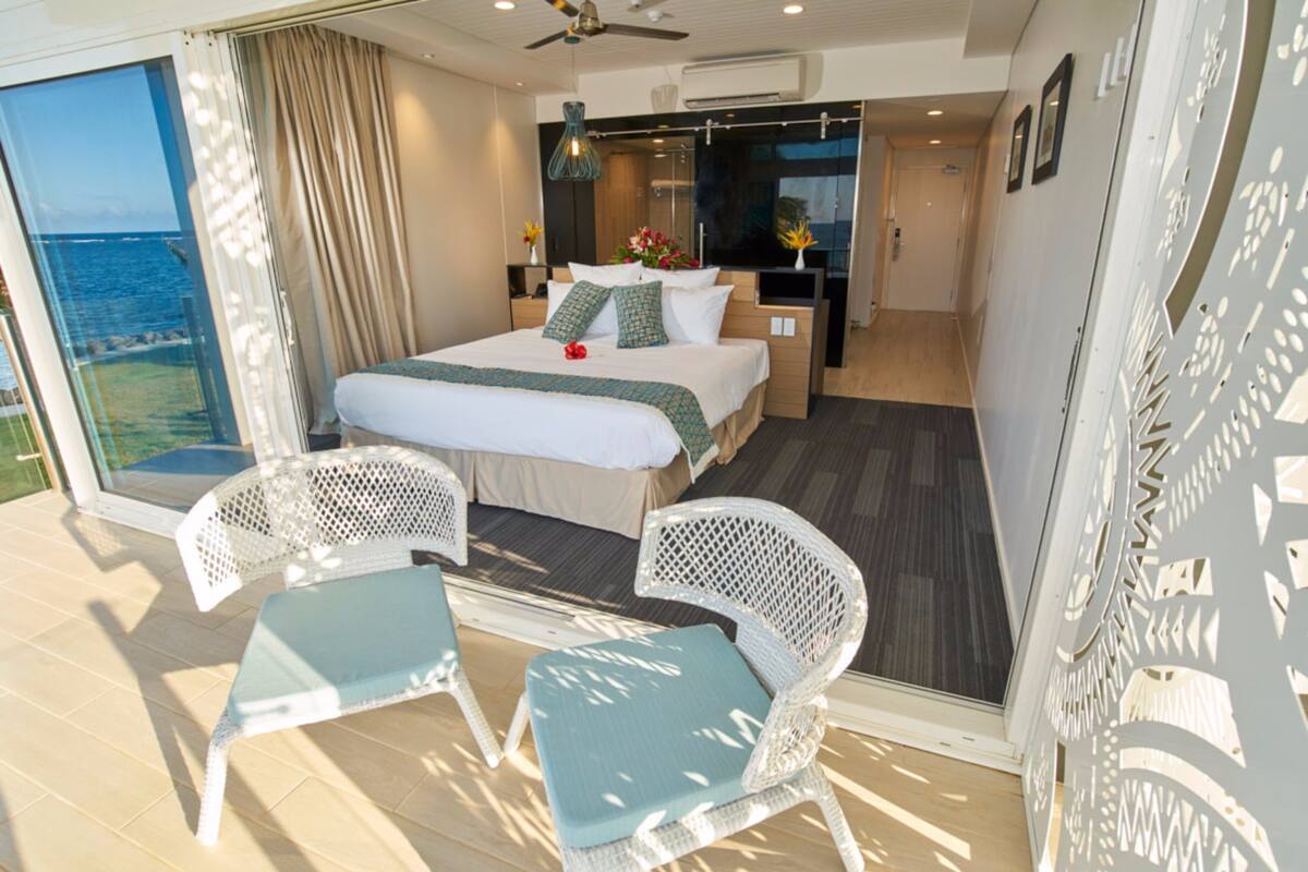 A hotel room with a bed, two white wicker chairs, a small table, a balcony with a view of the sea, and a ceiling fan.