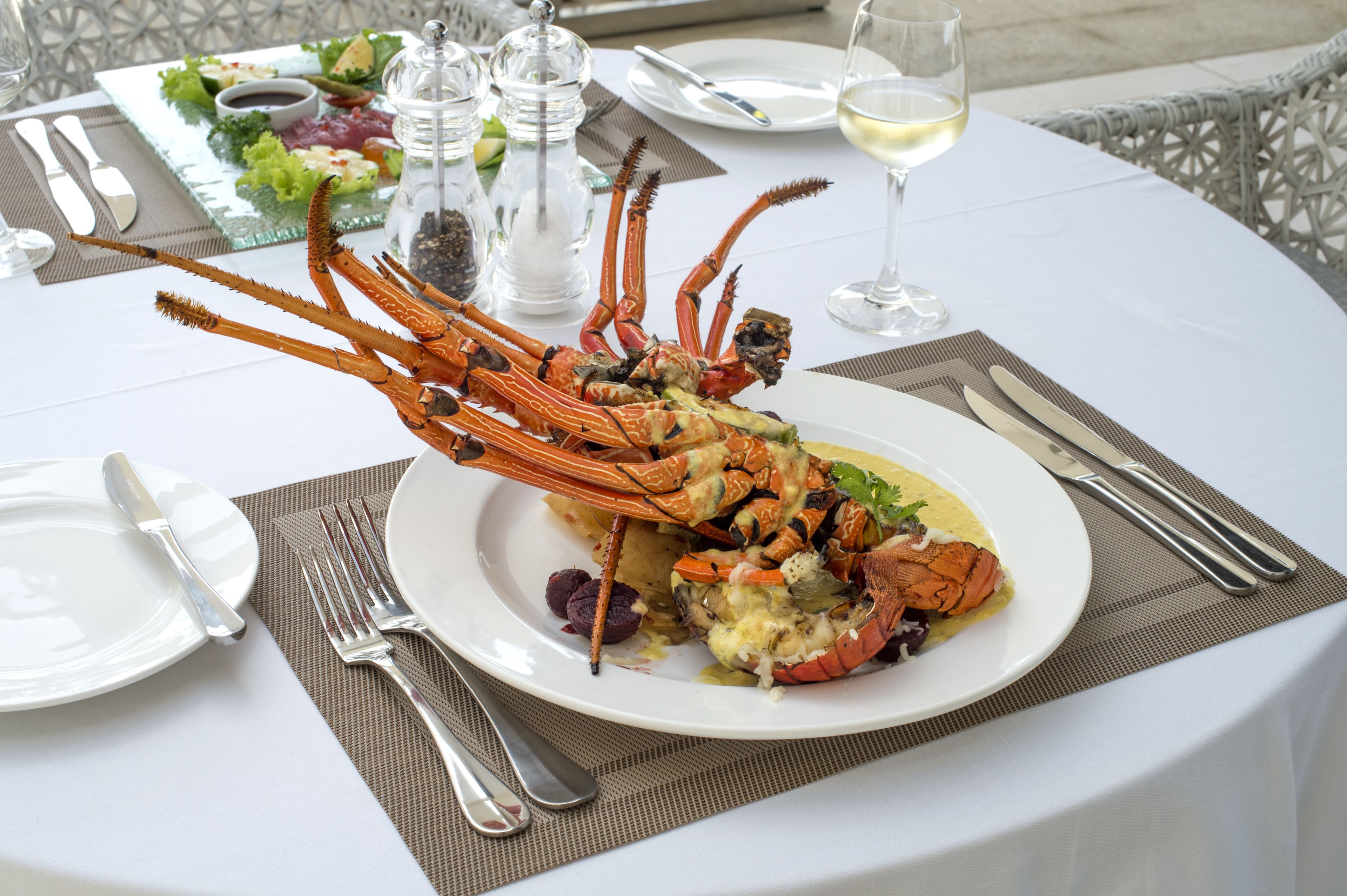 A lobster dish on a white plate with a fork and knife beside it, a glass of white wine, and a salad on a separate plate.