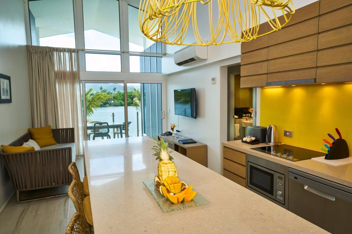 A modern kitchen with a central island, a dining area with a sofa, and a large window offering a view of greenery.
